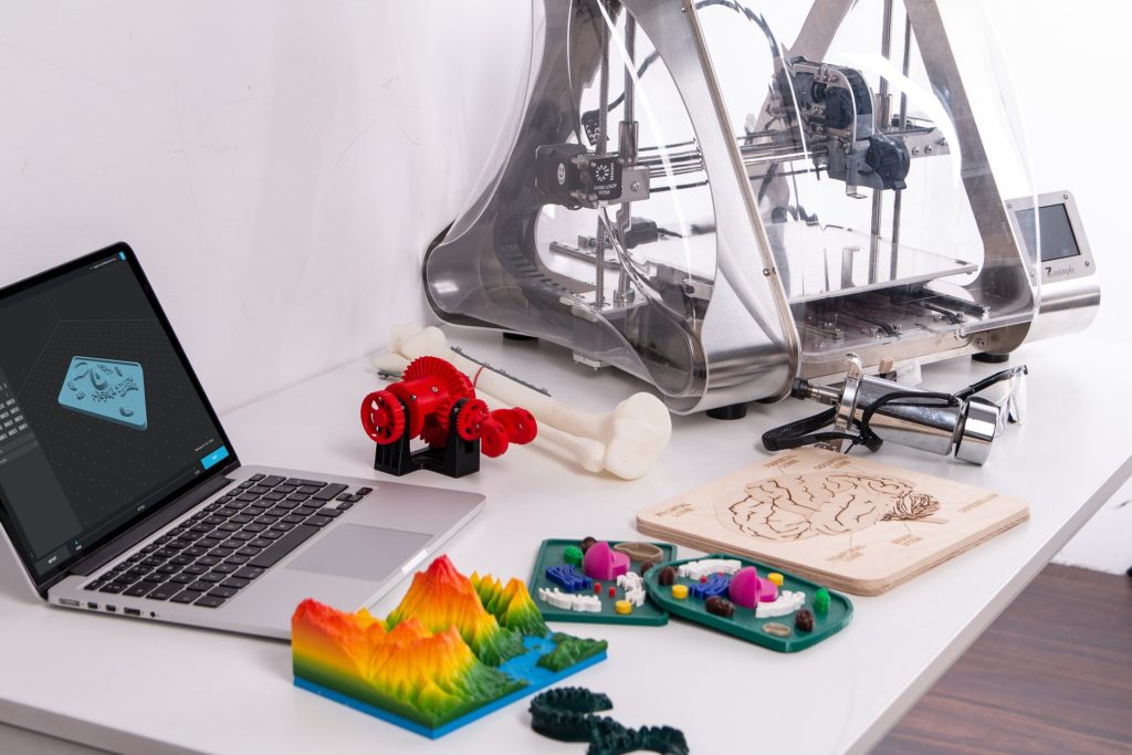 Computer beside a 3D printer with a number of 3D models of gears, a mountain range, bones, teeth, and dioramas.